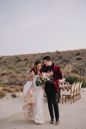 Bride and groom walking in desert landscape, she holds floral bouquet in lace gown, he wears burgundy velvet blazer.