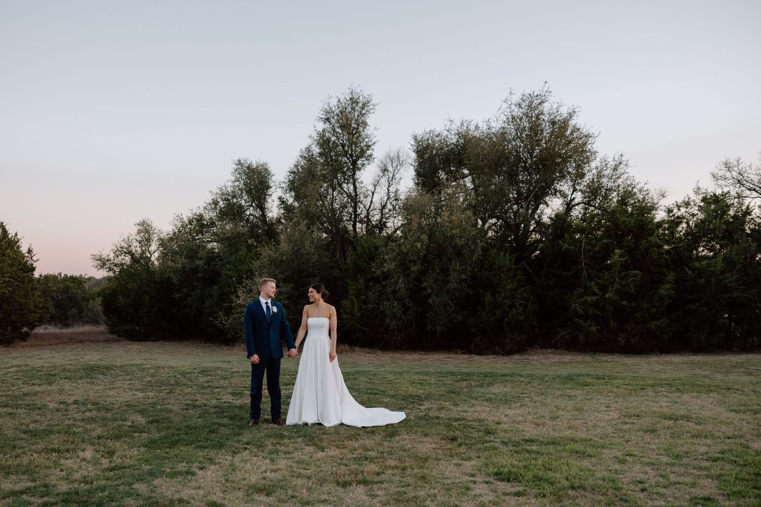 A couple stands together in an open field at dusk with trees silhouetted in the background.