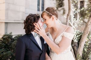 A romantic wedding couple embraces outdoors on a sunny day with greenery and twinkle lights in the background.