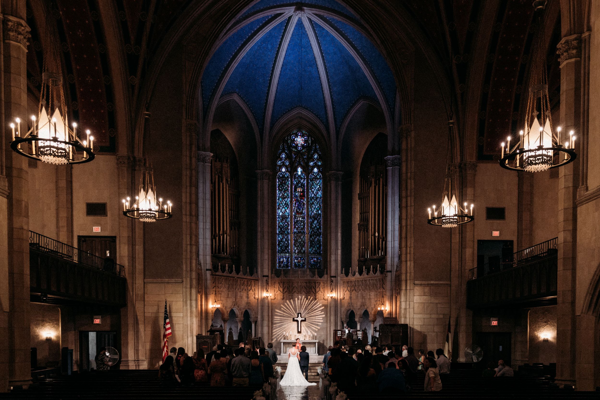 A wedding ceremony takes place in a grand cathedral with ornate architecture and glowing chandeliers illuminating the space.