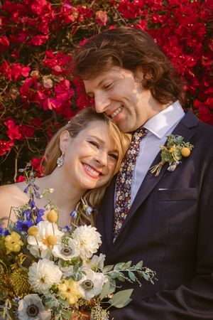 A romantic wedding couple embraces against a vibrant backdrop of red bougainvillea flowers in an intimate garden setting.