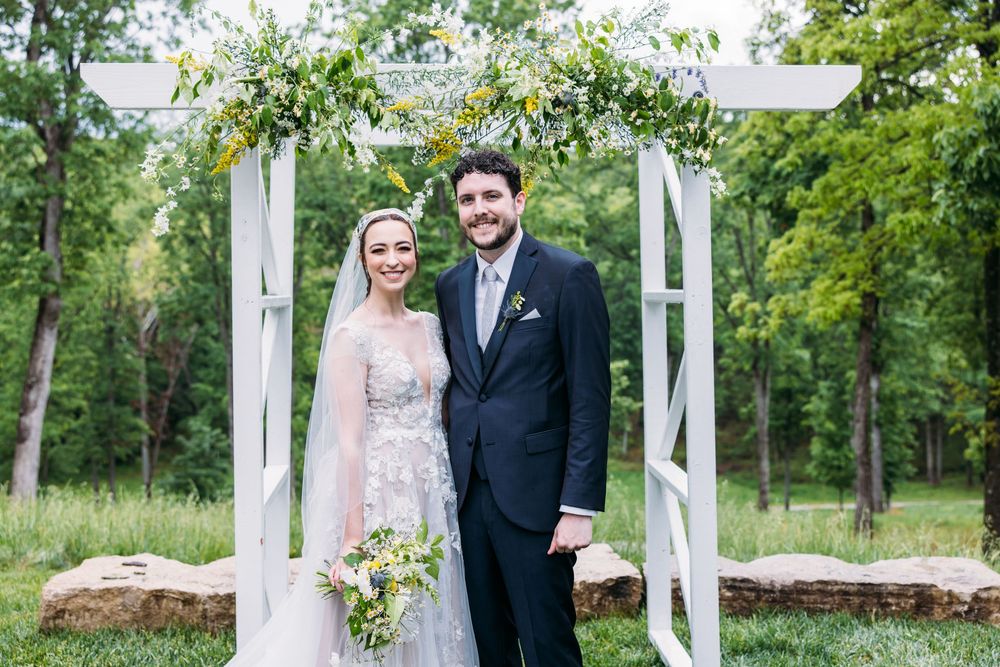 A couple poses together in front of a white pergola surrounded by lush green trees and natural scenery.