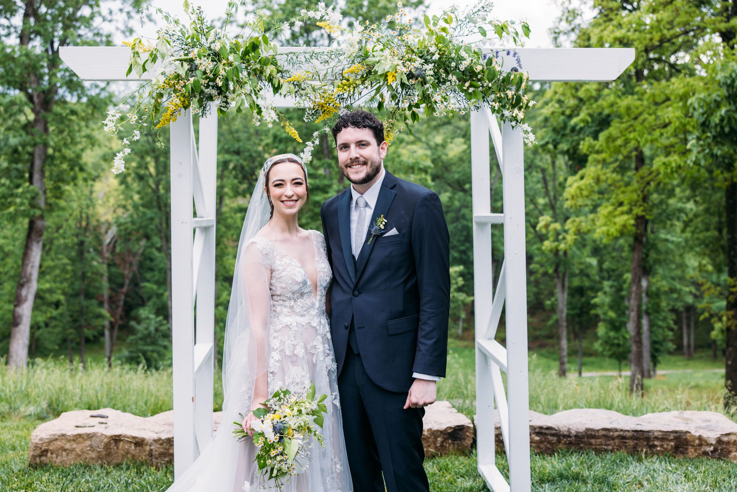 A couple poses together in front of a white pergola surrounded by lush green trees and natural scenery.