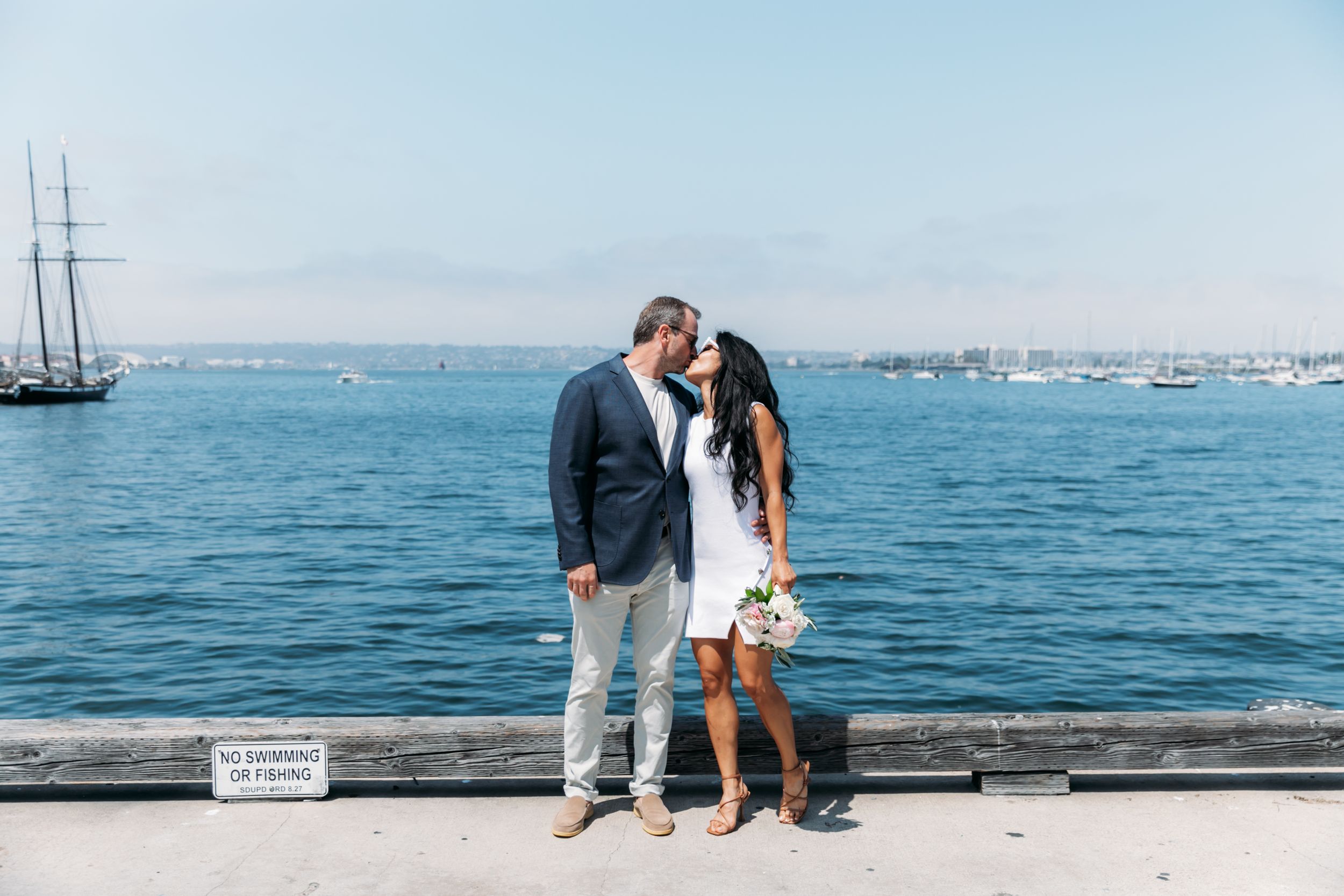 A couple shares a romantic moment on a pier overlooking the ocean with boats and a cityscape in the distance.
