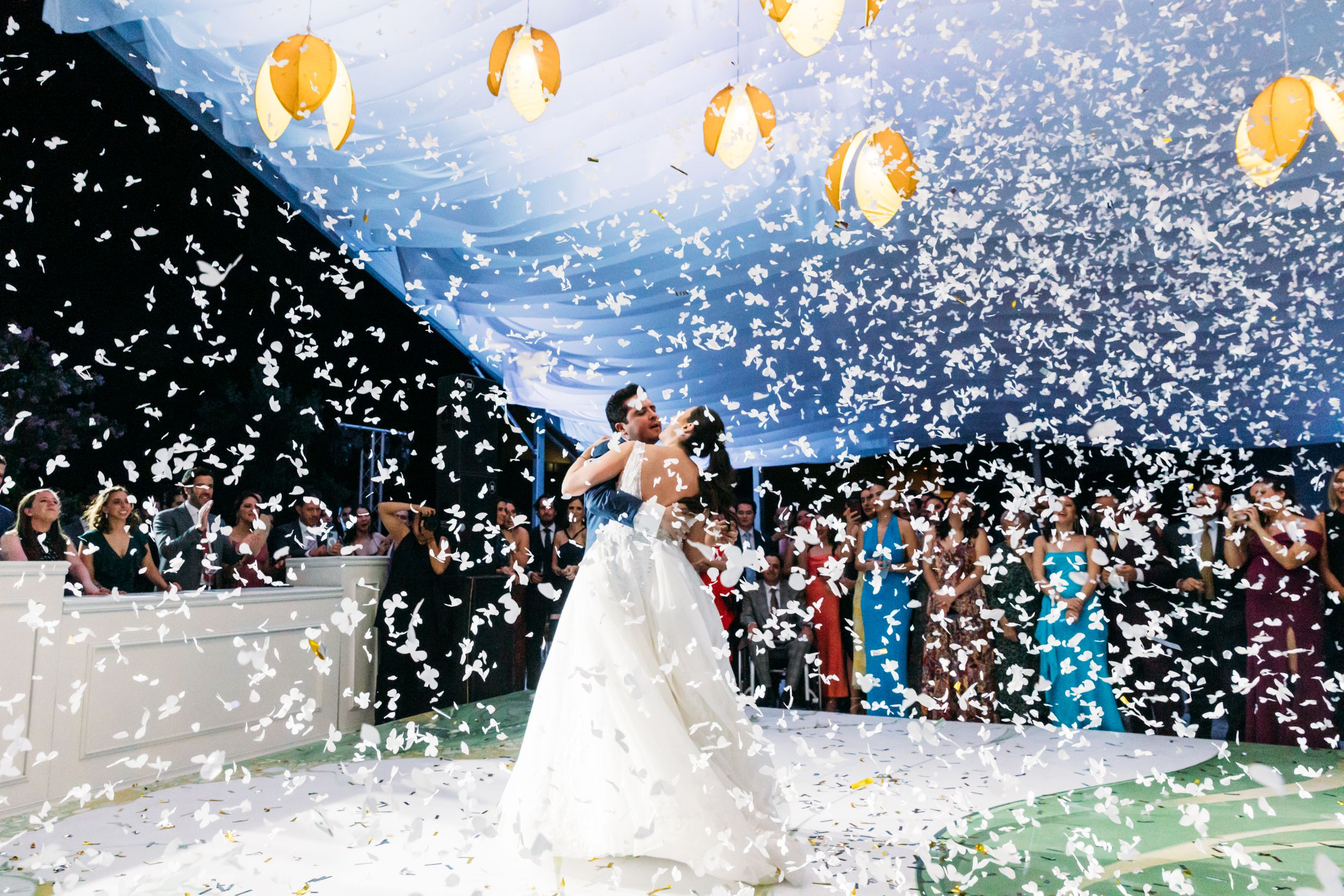A couple dances under glowing lanterns while confetti falls during an outdoor wedding celebration.