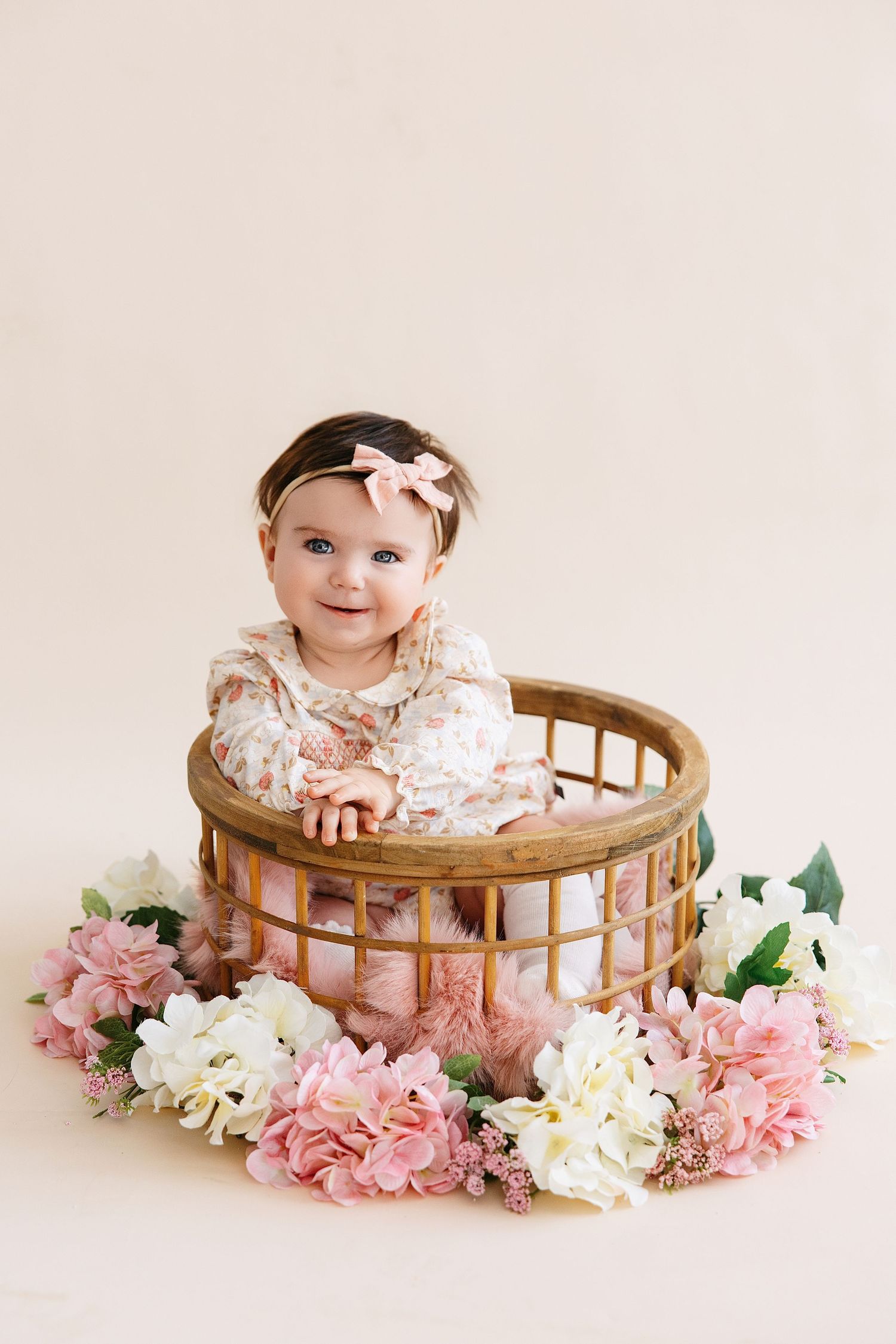 A baby sits in a round wooden basket surrounded by pink and white flowers in a soft studio setting.