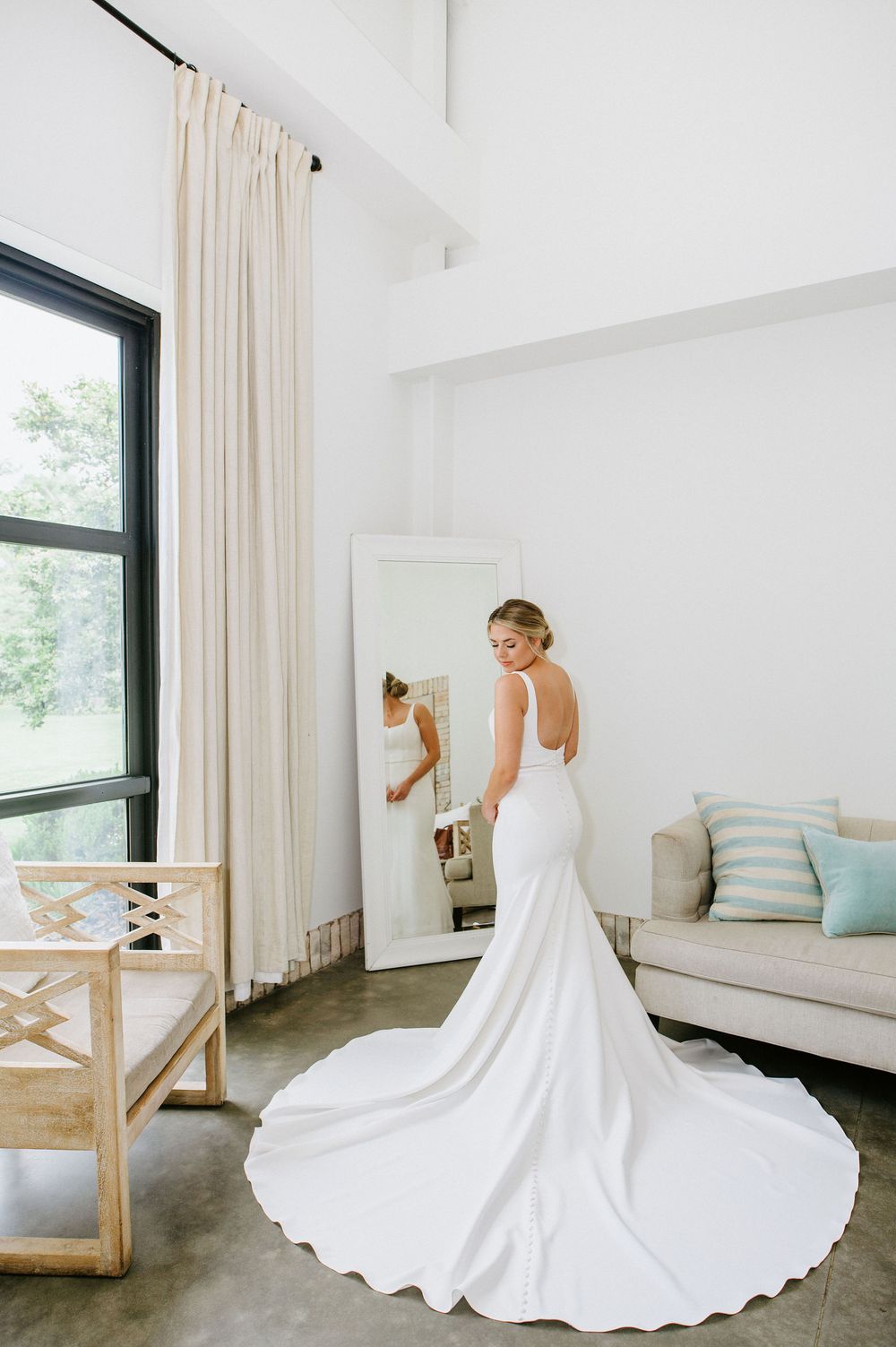 A flowing white wedding dress with a dramatic train spreads elegantly across a modern minimalist room with cream curtains.