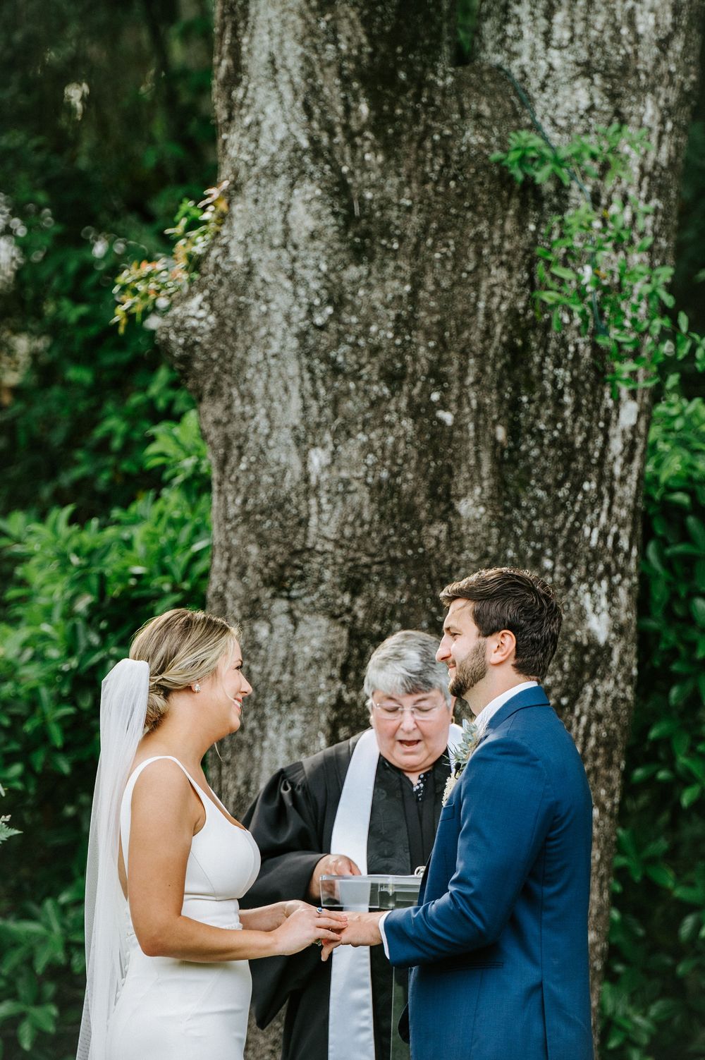 A wedding ceremony takes place under majestic oak trees with Spanish moss as guests look on.