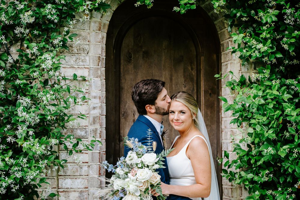 A couple shares an intimate moment in a garden archway surrounded by lush greenery and stone architecture.