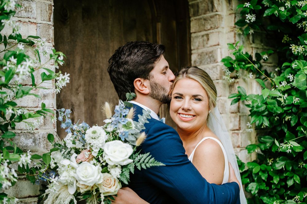 A couple in wedding attire embraces near a stone wall covered in lush green vines and white floral arrangements.