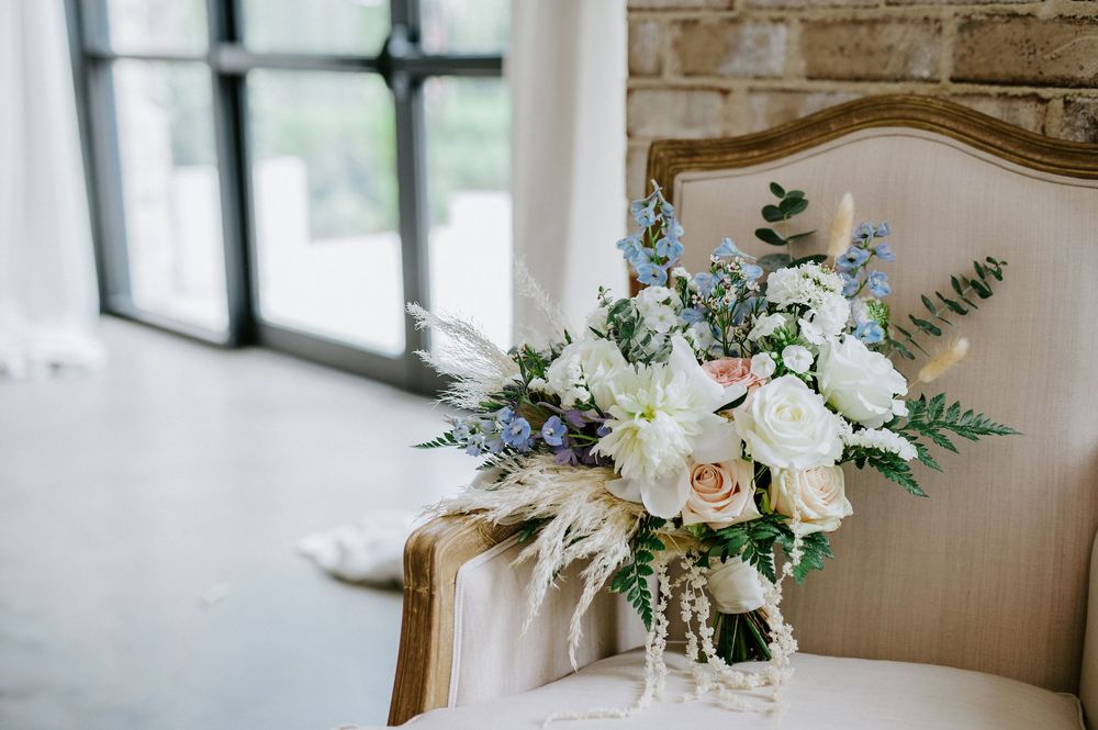 White and blue floral wedding bouquet with eucalyptus leaves rests on rustic wooden table near brick wall.
