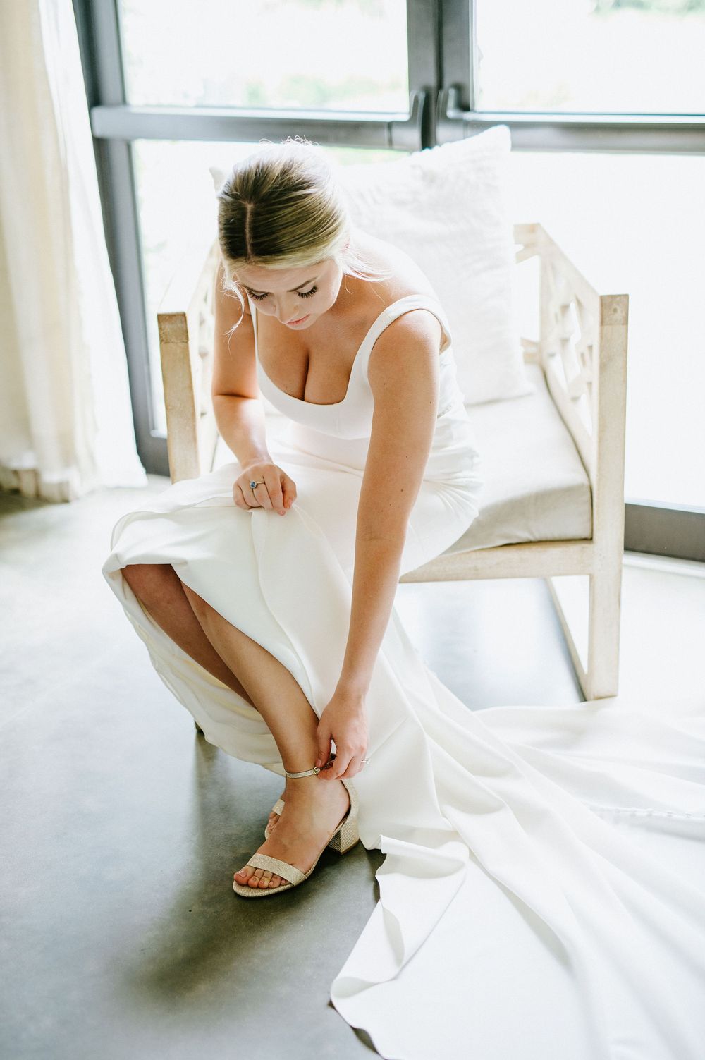 Someone in a white wedding dress adjusts their sandal while sitting in a bright sunlit room.