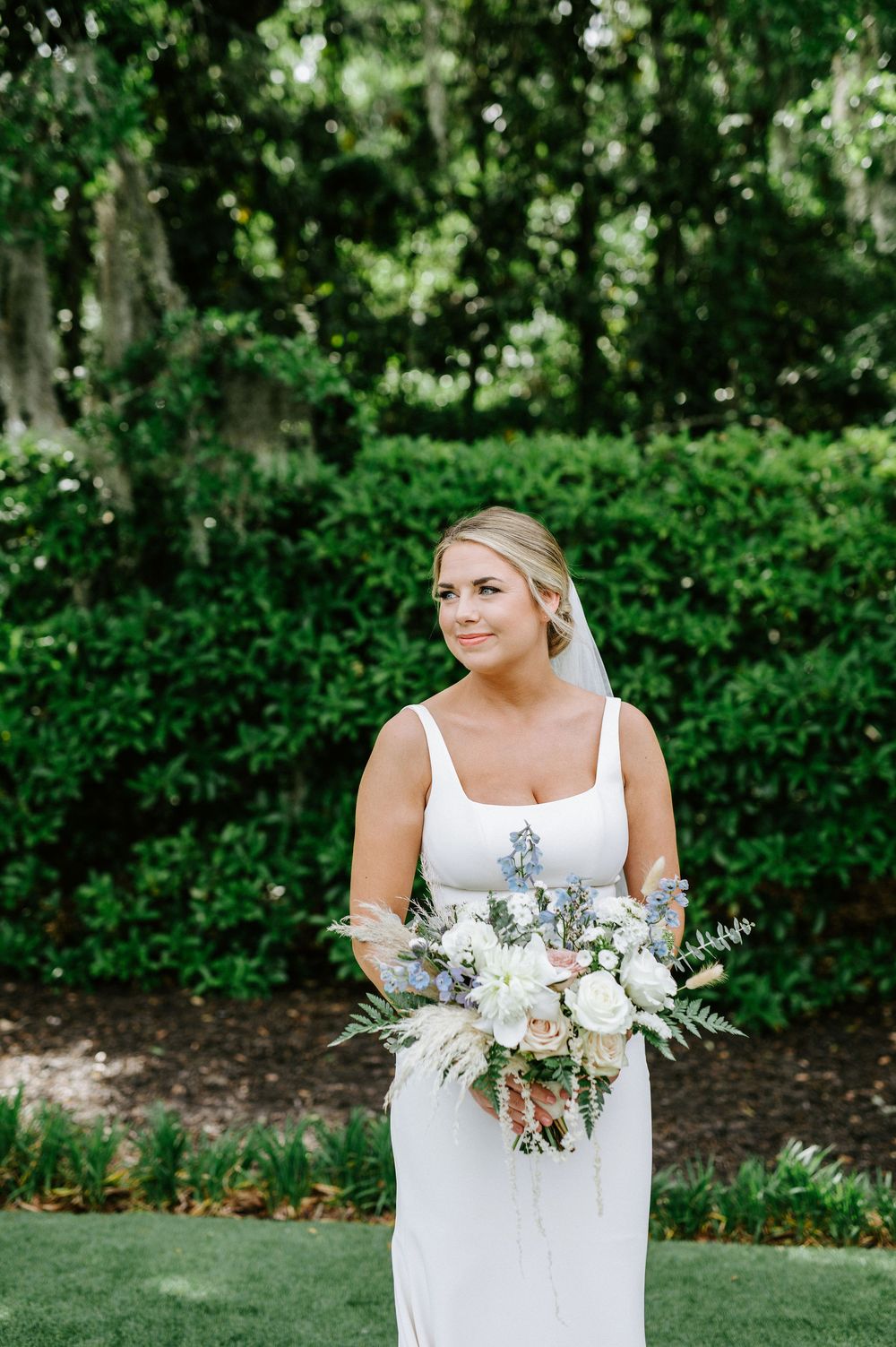 A figure in a white wedding dress holds a delicate bouquet of white and blue flowers against a lush garden backdrop.