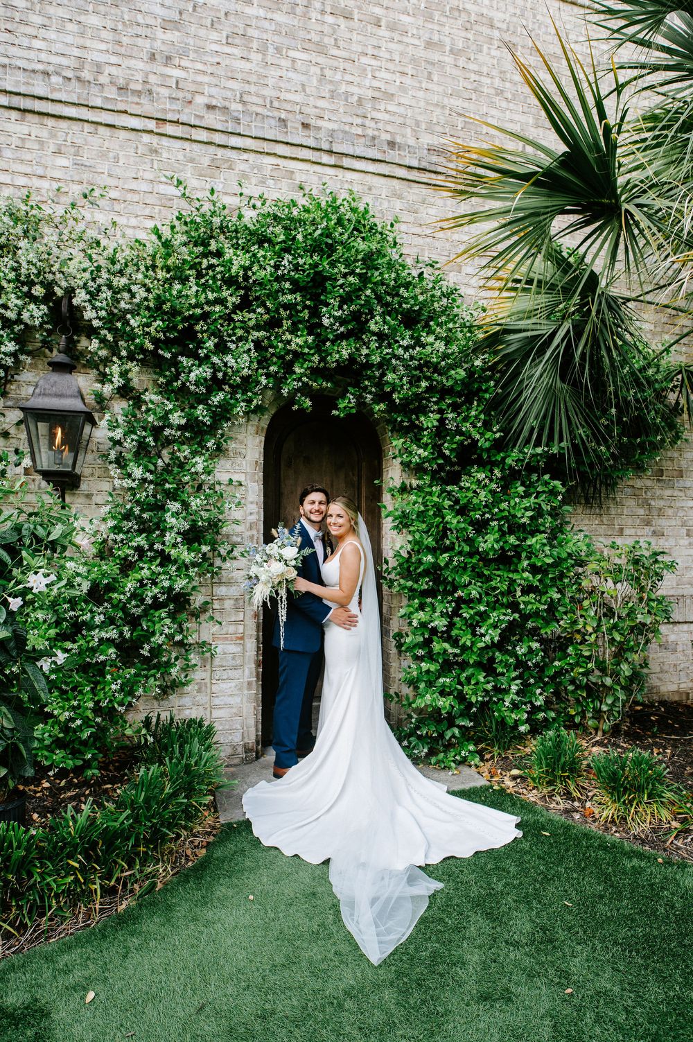 A white wedding dress flows elegantly against a lush green garden wall with brick backdrop in a romantic outdoor setting.