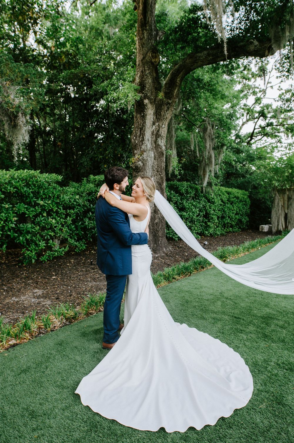 A couple in wedding attire dances gracefully on a lush green lawn surrounded by trees and foliage.