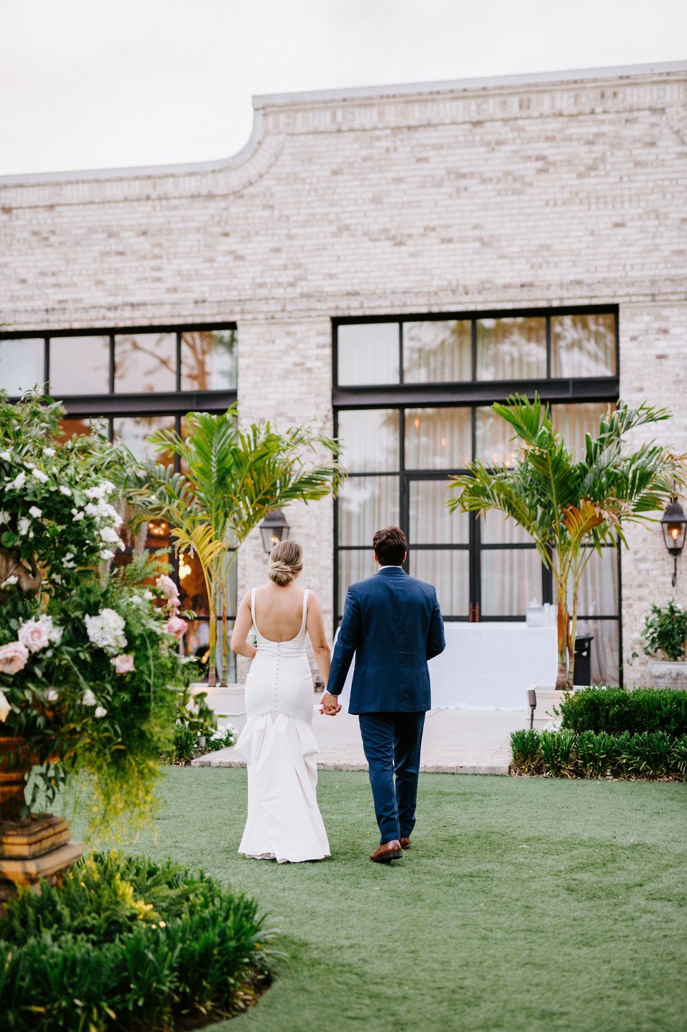 A couple walks together on a manicured lawn towards a white brick building with palm trees and greenery.