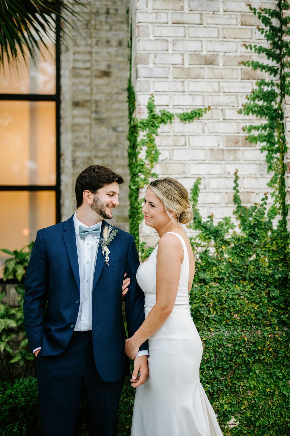 A couple shares joyful moments together against a white brick wall and greenery on their wedding day.