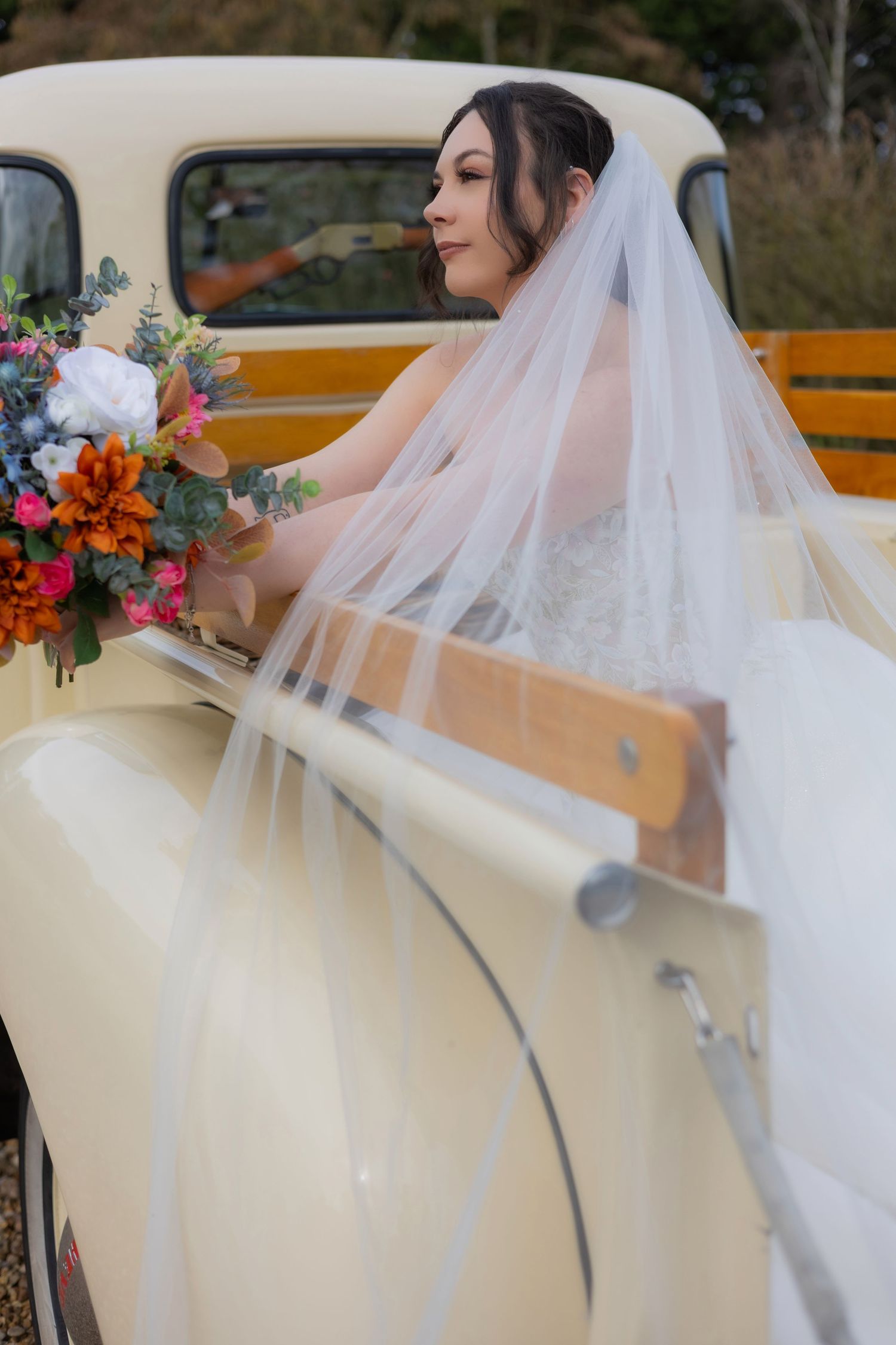 Bride in white dress and veil seated in vintage cream-colored car.