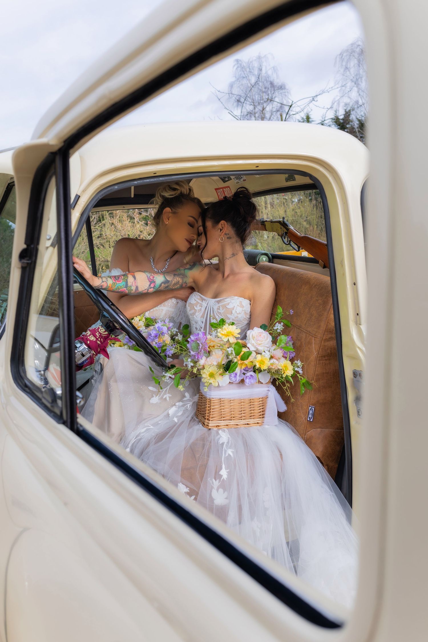Romantic moment captured through vintage car window with white wedding dresses and floral bouquet.