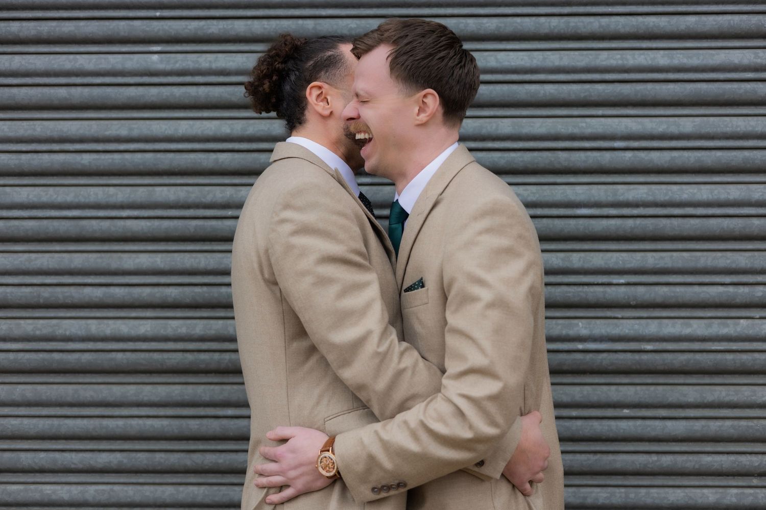 Couple in matching beige outfits embracing against an industrial metal backdrop.