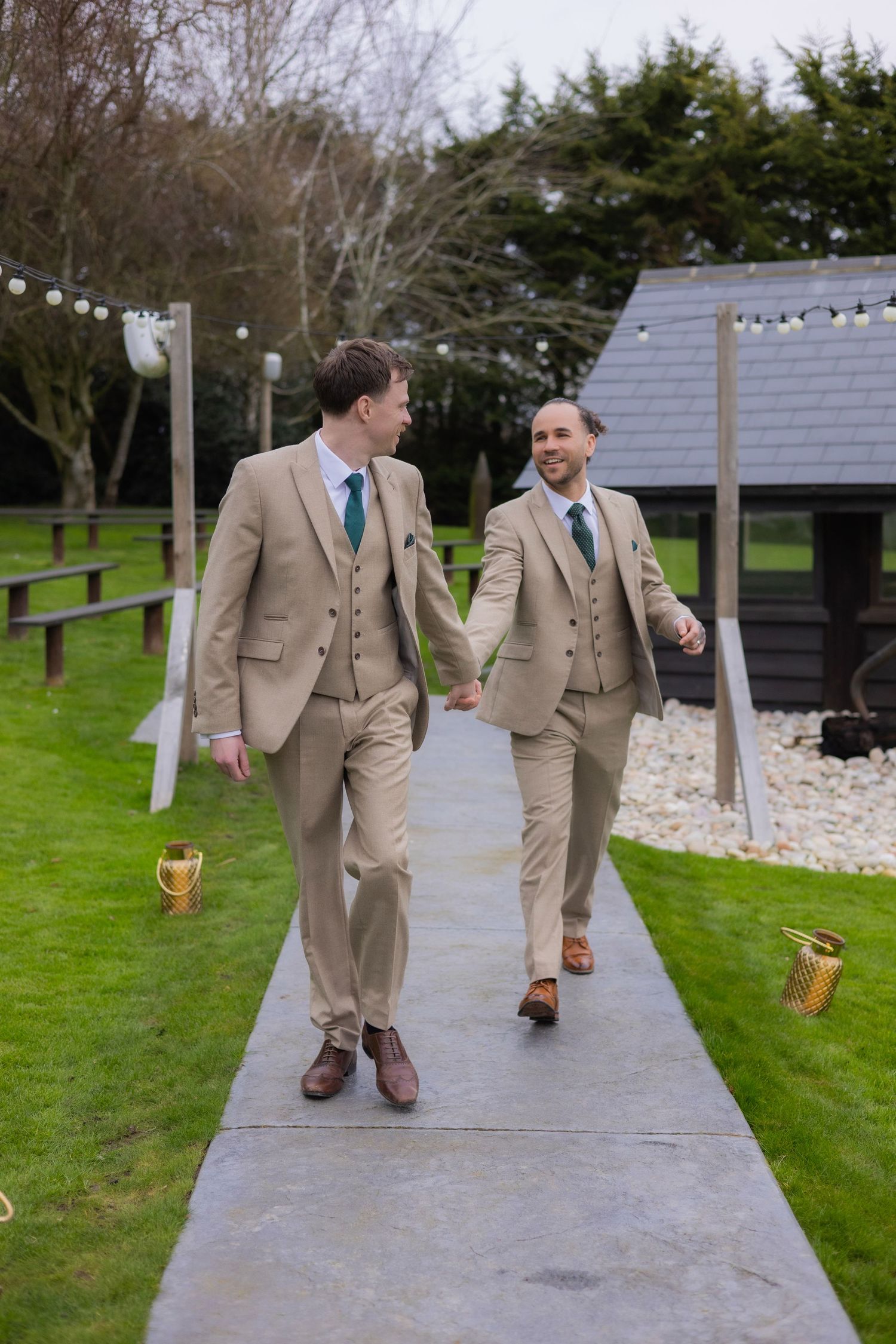 Two people in matching beige suits walk joyfully down a path with string lights overhead.