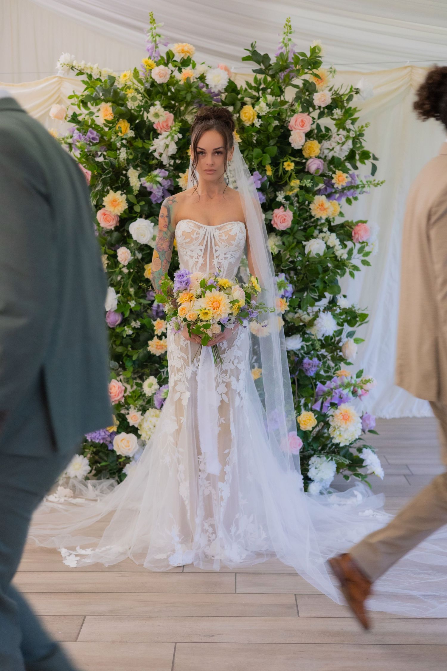 Bride in lace gown stands under floral arch with yellow and purple blooms.