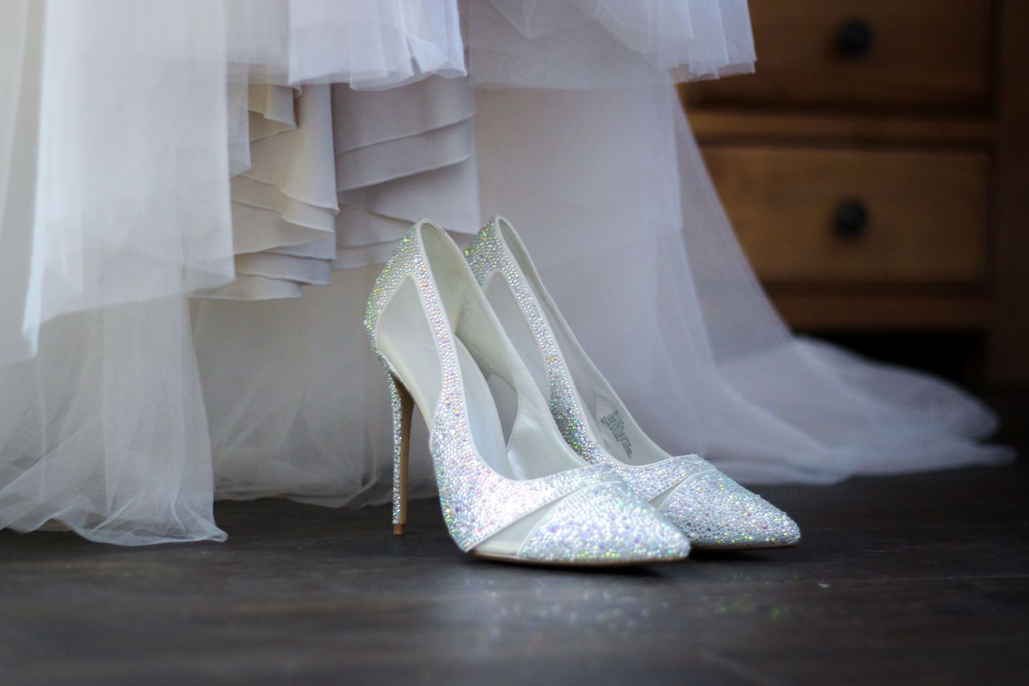 Close-up of white bridal shoes and dress hem on wooden floor.