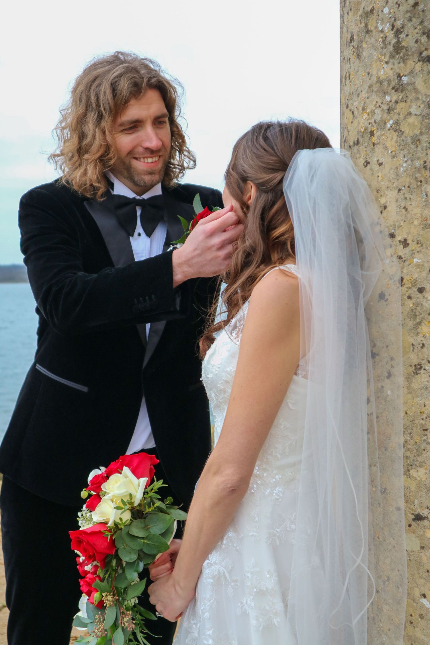 Intimate wedding moment captured by the sea with red and white bouquet.