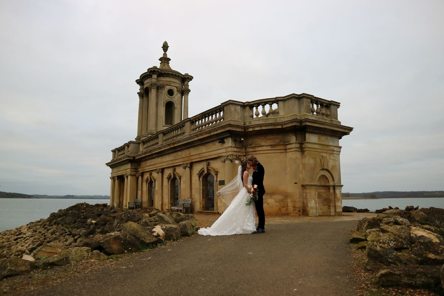 Historic stone chapel overlooking water creates dramatic backdrop for wedding photography.