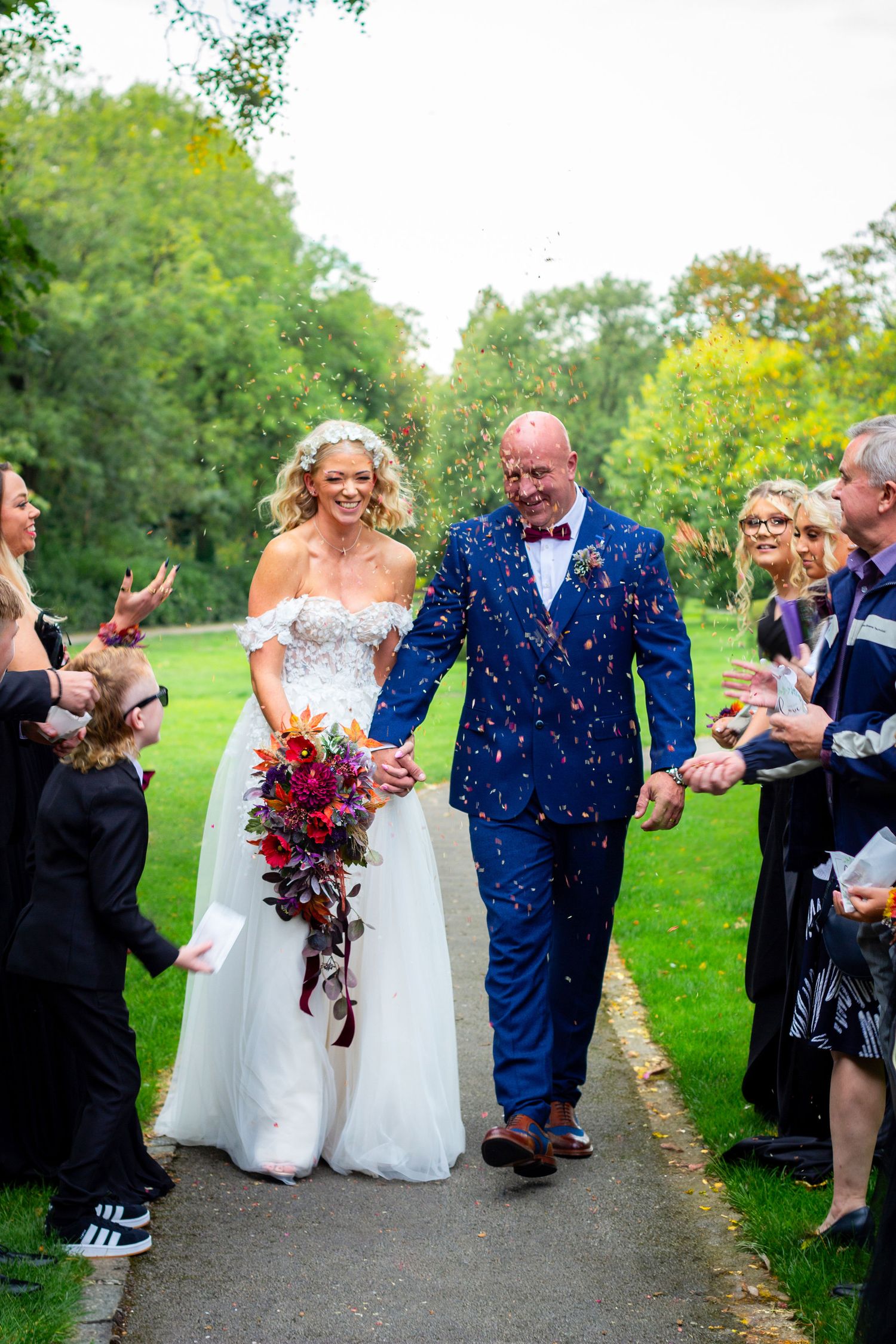 Wedding couple walks through shower of confetti on path lined with celebrating guests.