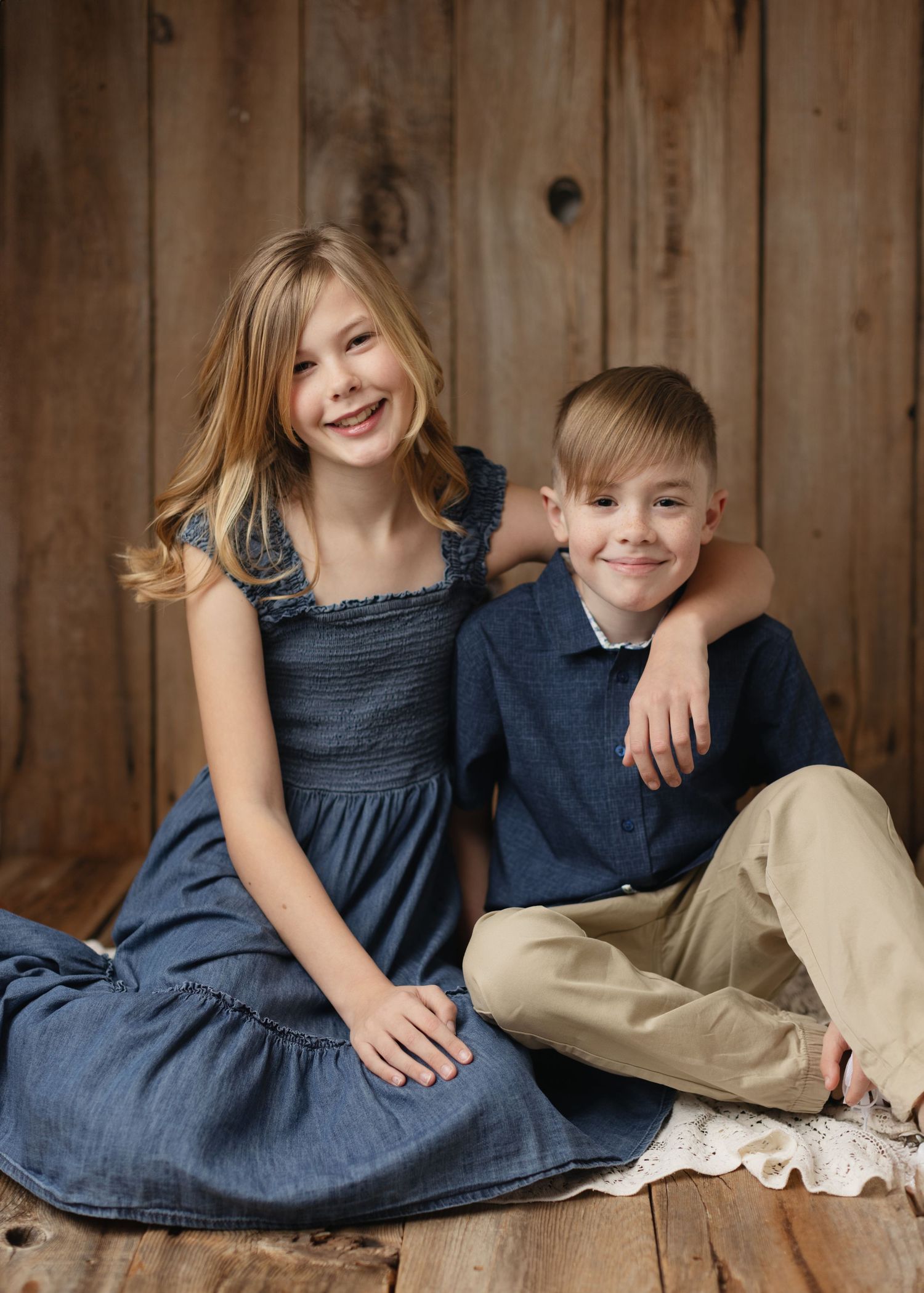 Two children in coordinating blue and khaki outfits pose together against a rustic wooden wall background. Elizabeth Lang Photography Surrey