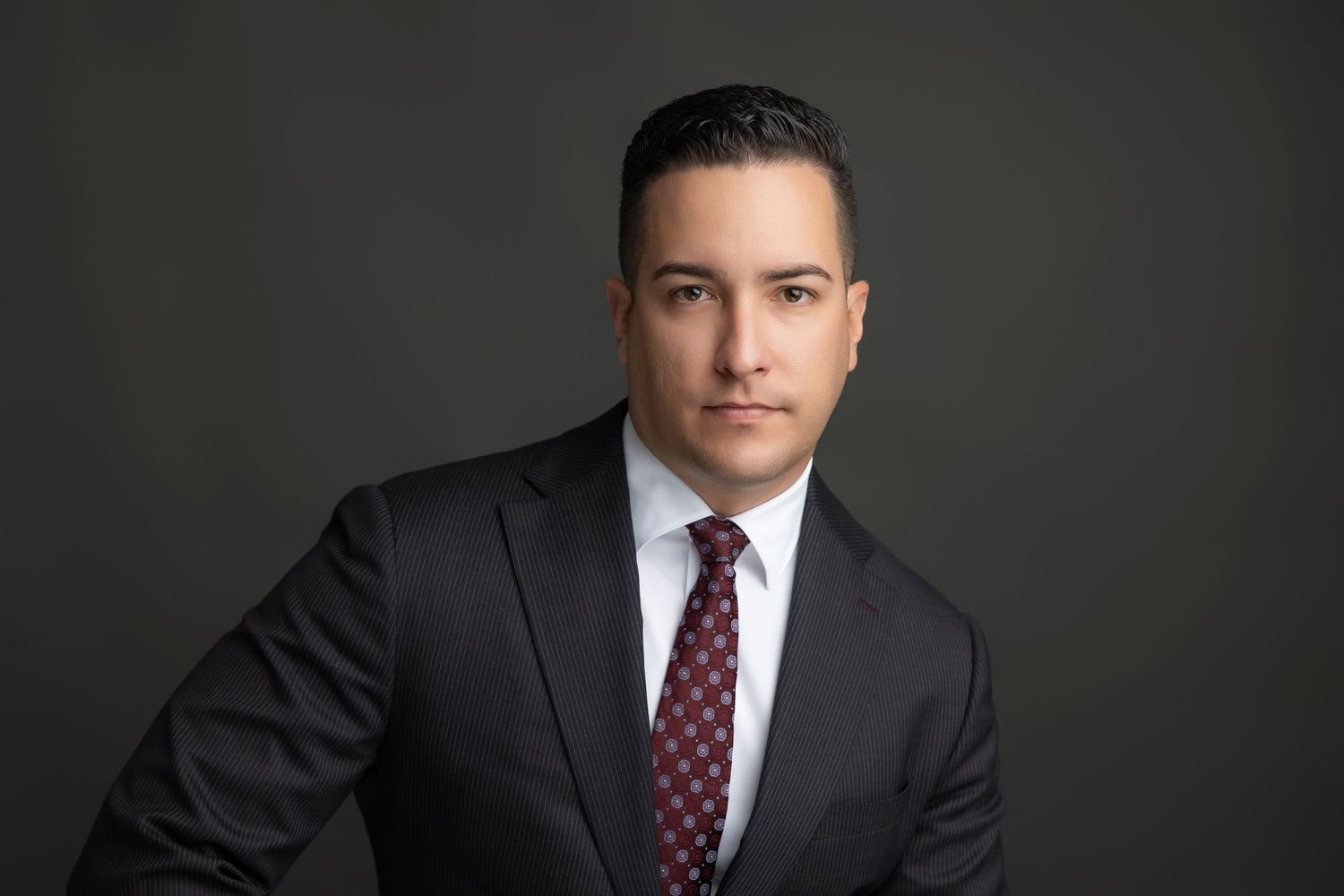 Professional headshot portrait of a person in a dark suit and burgundy polka dot tie against a gray background.
