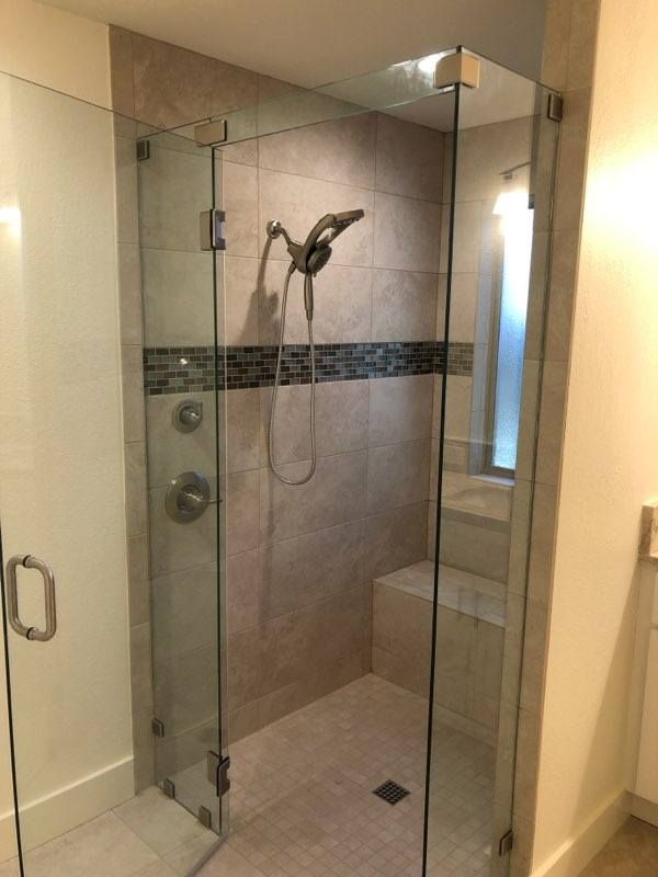 Modern glass shower enclosure with frameless doors, mosaic tile accent strip, and handheld showerhead in beige bathroom.