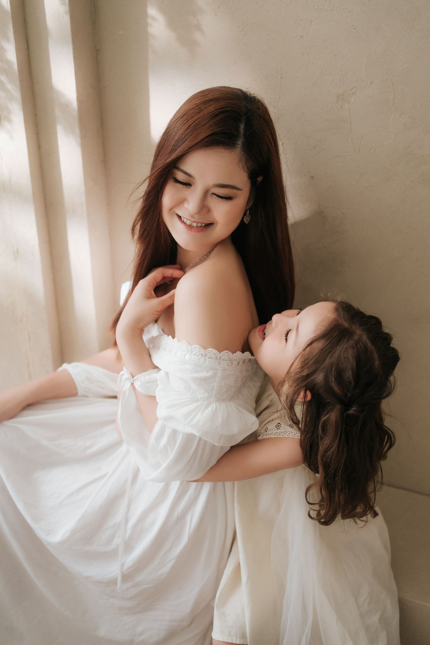 A smiling young woman in a white off-shoulder dress tenderly holds a little girl in white, both with long dark hair.一位微笑的年輕女子身穿白色露肩連身裙
