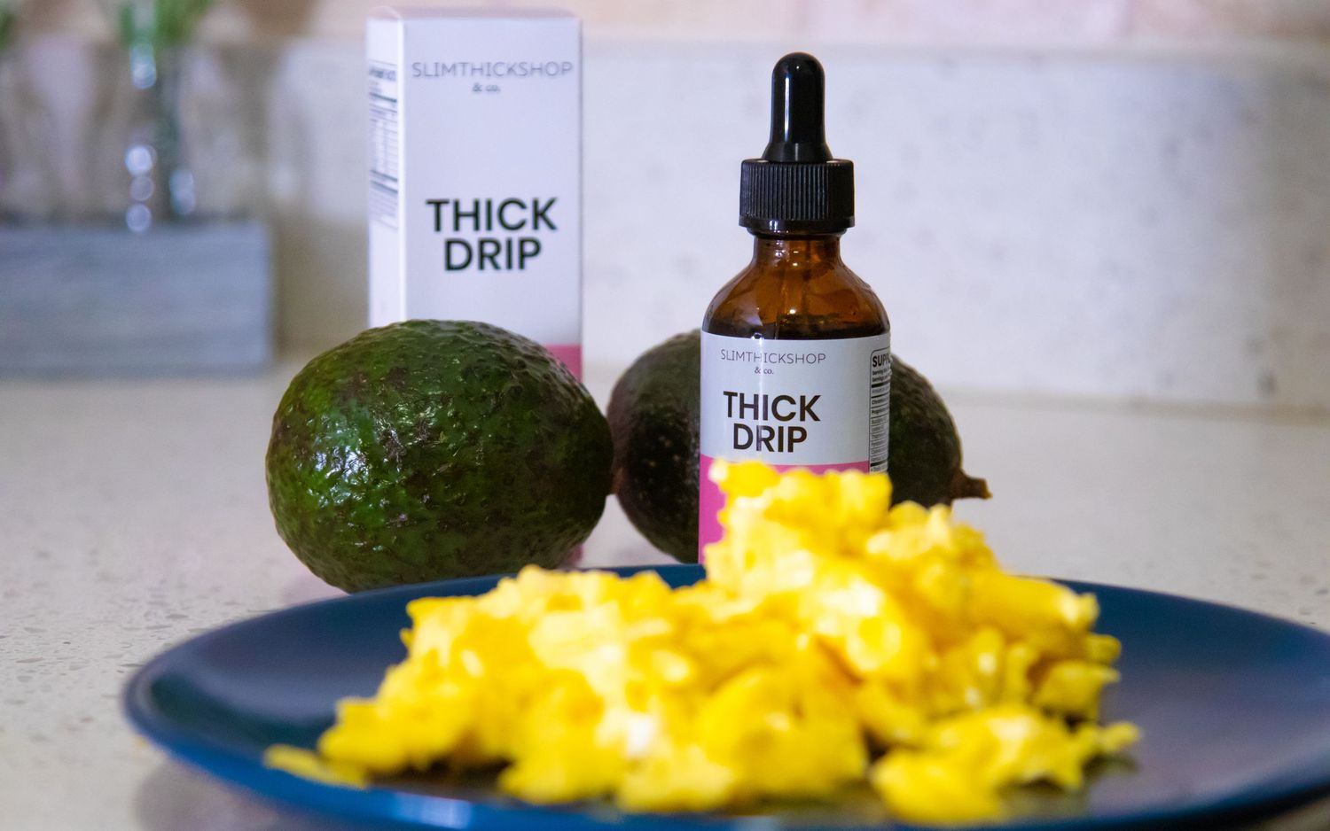 Thick Drip hair oil dropper bottle displayed with avocados and scrambled eggs on kitchen counter.