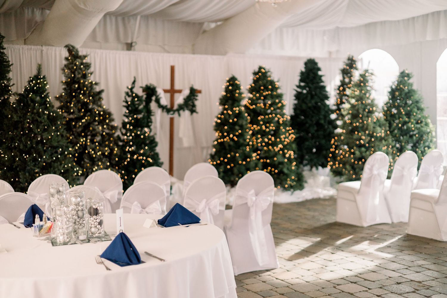 A white winter wonderland event space with decorated Christmas trees and elegant table settings under a draped tent ceiling.
