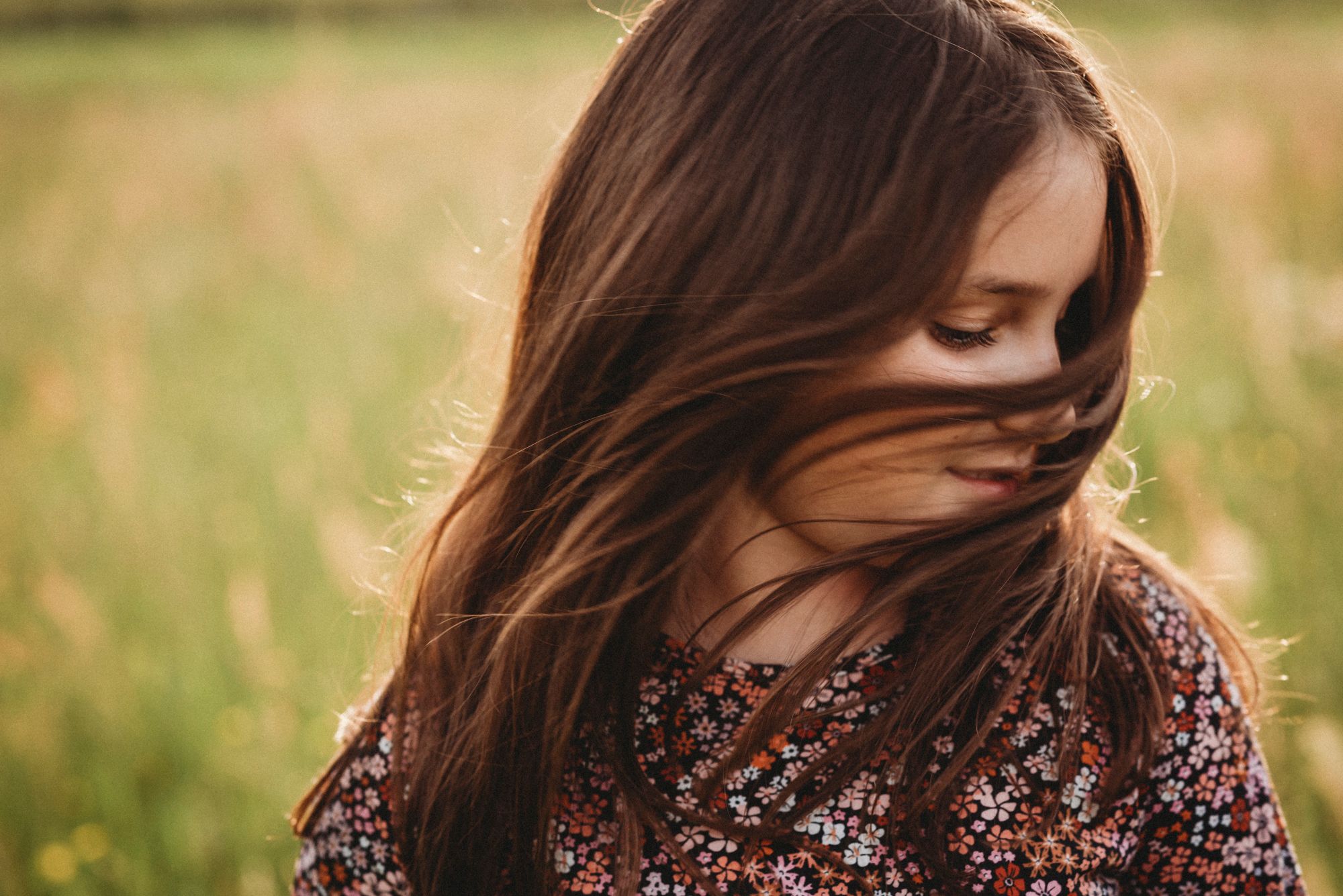 Person with long hair in floral dress stands in windswept meadow at golden hour.
