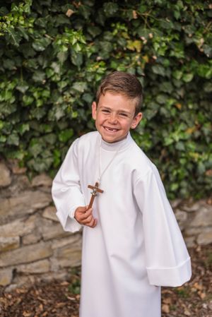 Person in white ceremonial robe holding cross stands before ivy-covered wall.