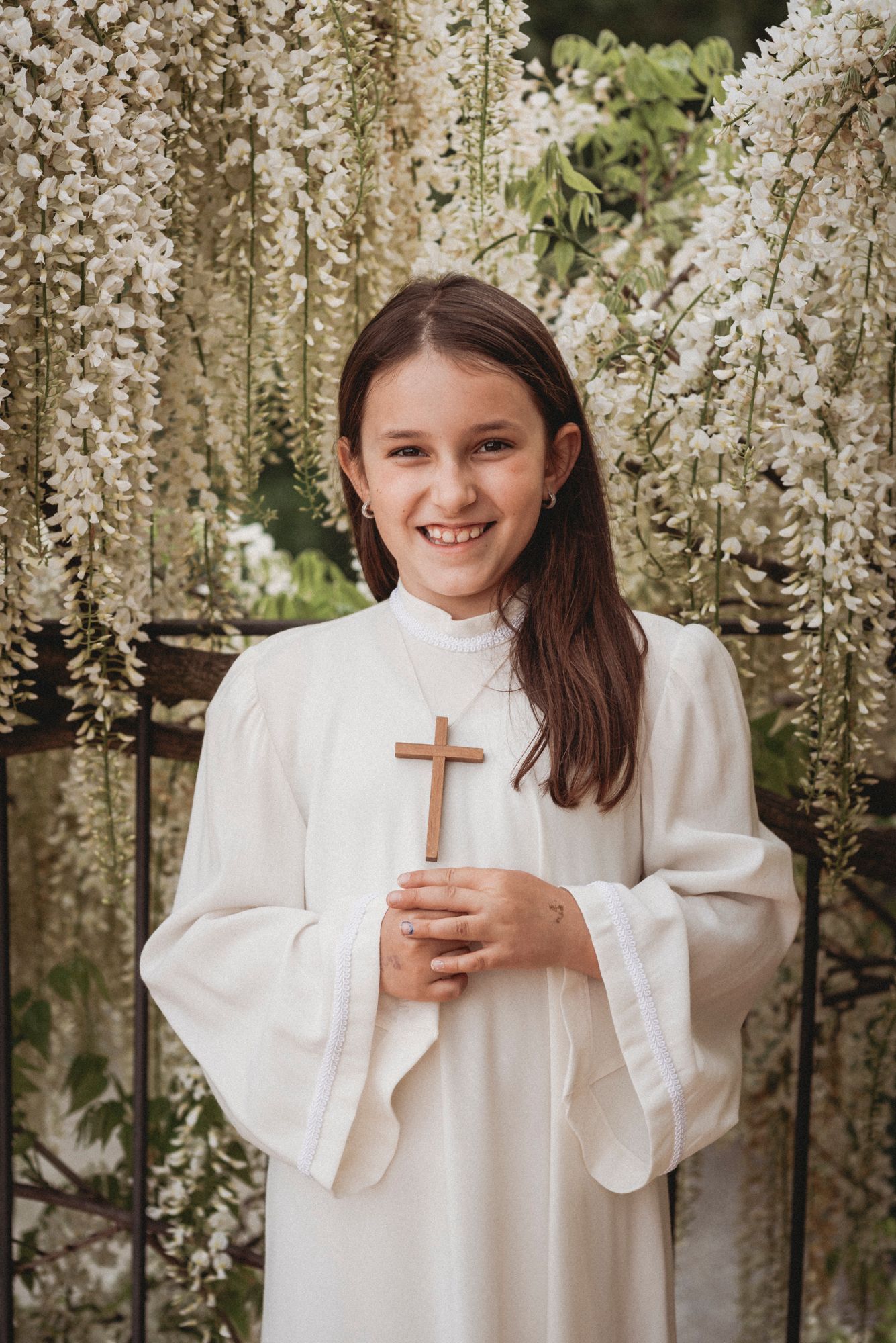 Person in white confirmation robe holding cross under cascading wisteria flowers.