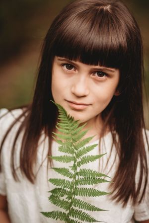 Close-up portrait with long dark bangs holding a green fern leaf against a blurred natural background.