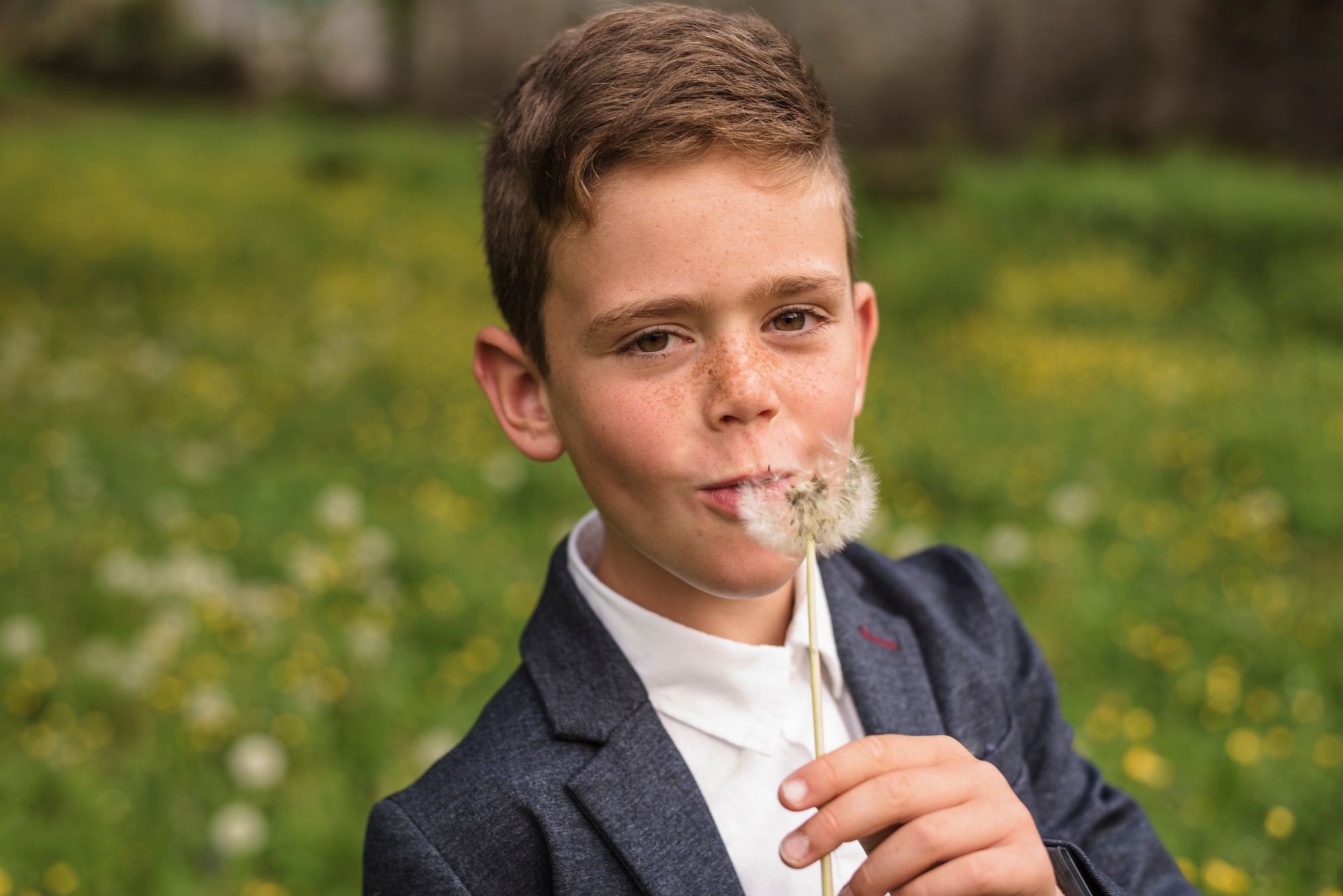 Child in navy blazer blowing on a dandelion in a spring meadow.