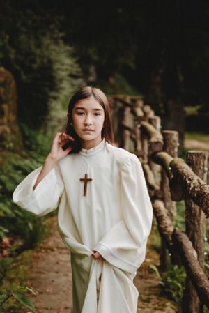Person in white ceremonial robe with cross standing on rustic woodland path.