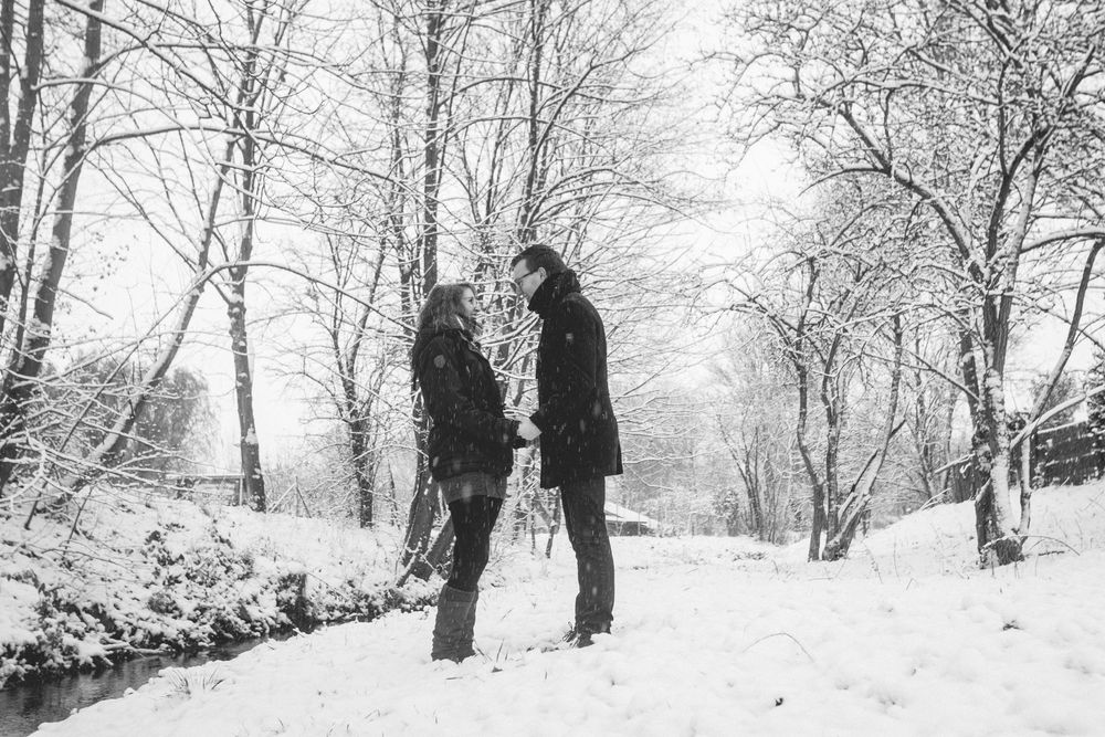 A couple stands together in a snowy winter landscape surrounded by bare trees in black and white.