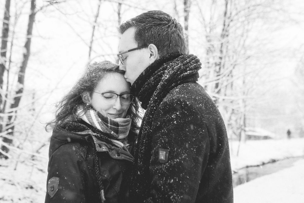 A couple embraces in a snowy winter forest setting captured in black and white photography.
