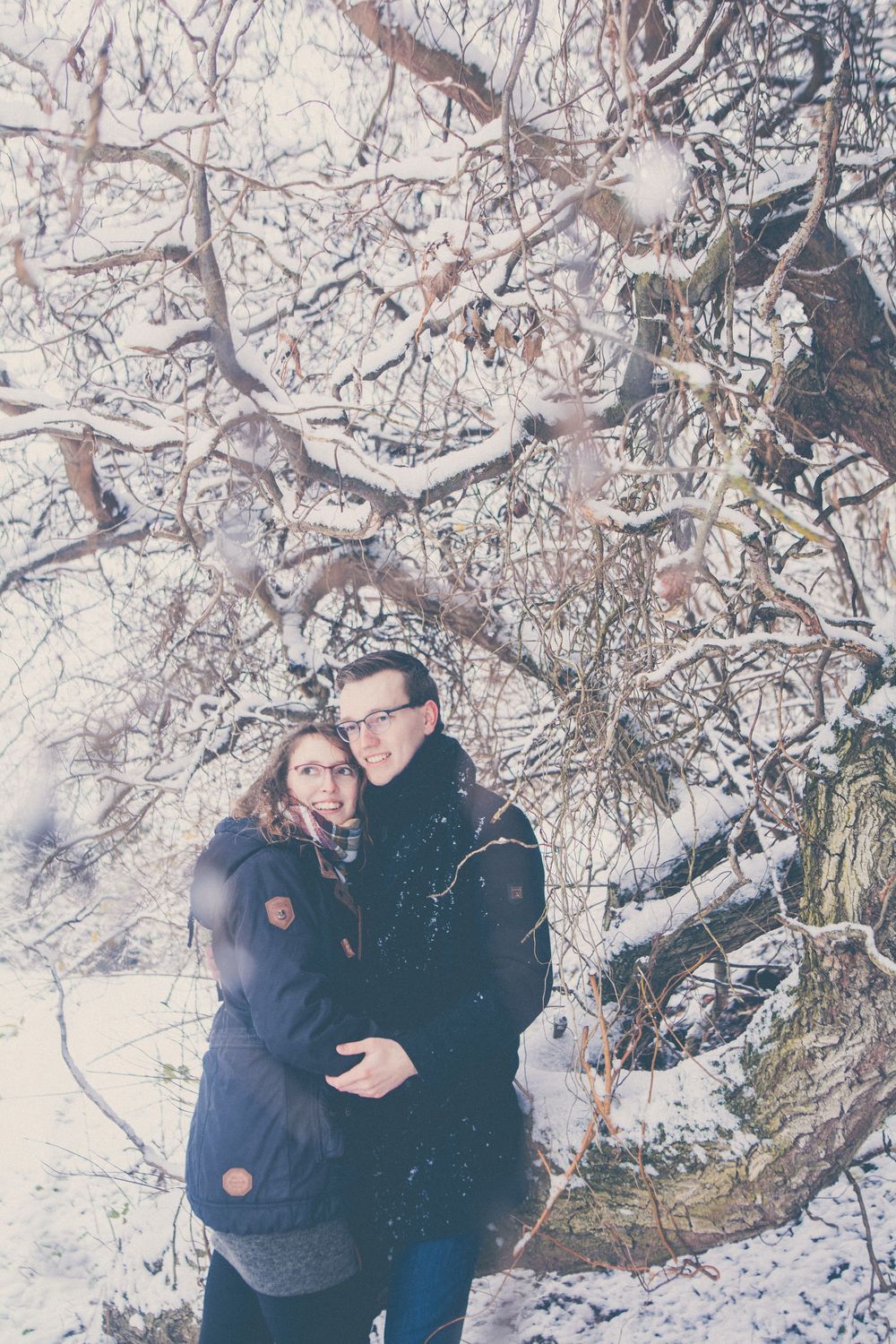 A couple shares romantic moments in a snowy winter landscape beneath frost-covered tree branches.