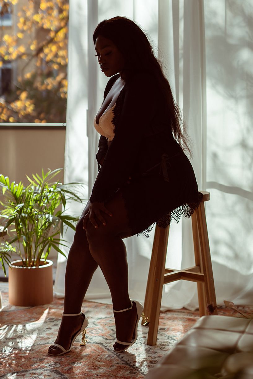Silhouette figure in black lingerie poses on wooden stool by window with potted plant and golden sunlight streaming in.