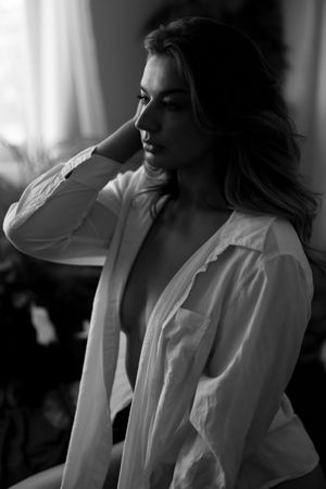 A dramatic black and white portrait showing a person in an oversized white shirt gazing to the side near a window.