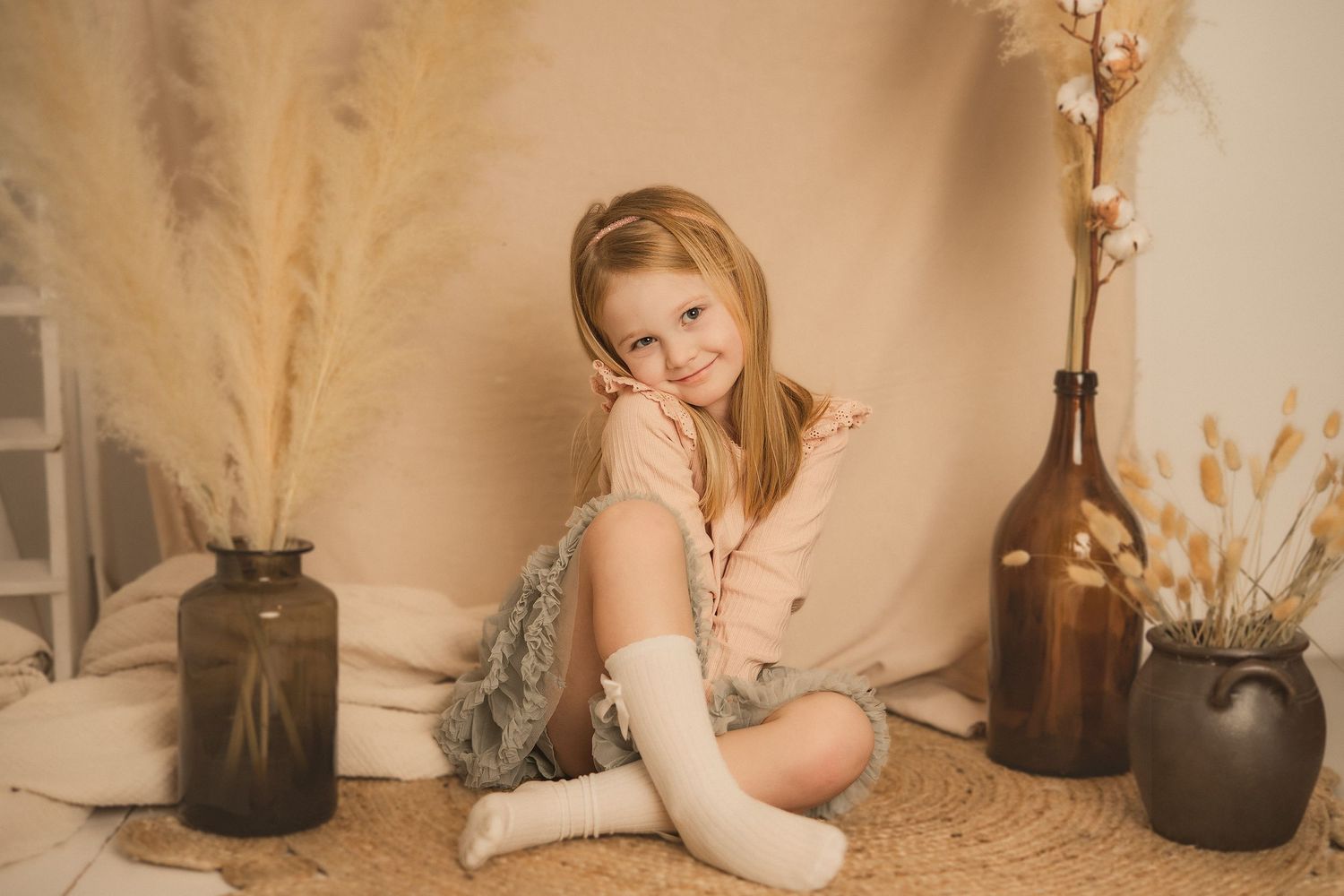A child sits on a beige backdrop surrounded by decorative vases and pampas grass in a warm, cozy setting.