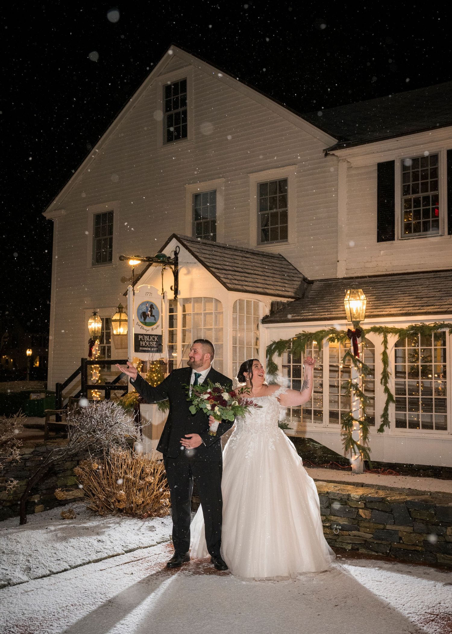 A couple shares romantic moments in the snow outside a white colonial-style venue with illuminated windows and lamp posts.