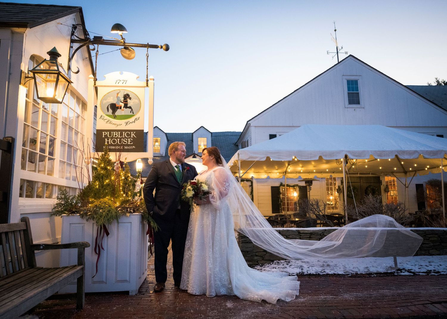 A wedding couple poses in front of a white farmhouse venue with snow-covered grounds on a winter day.